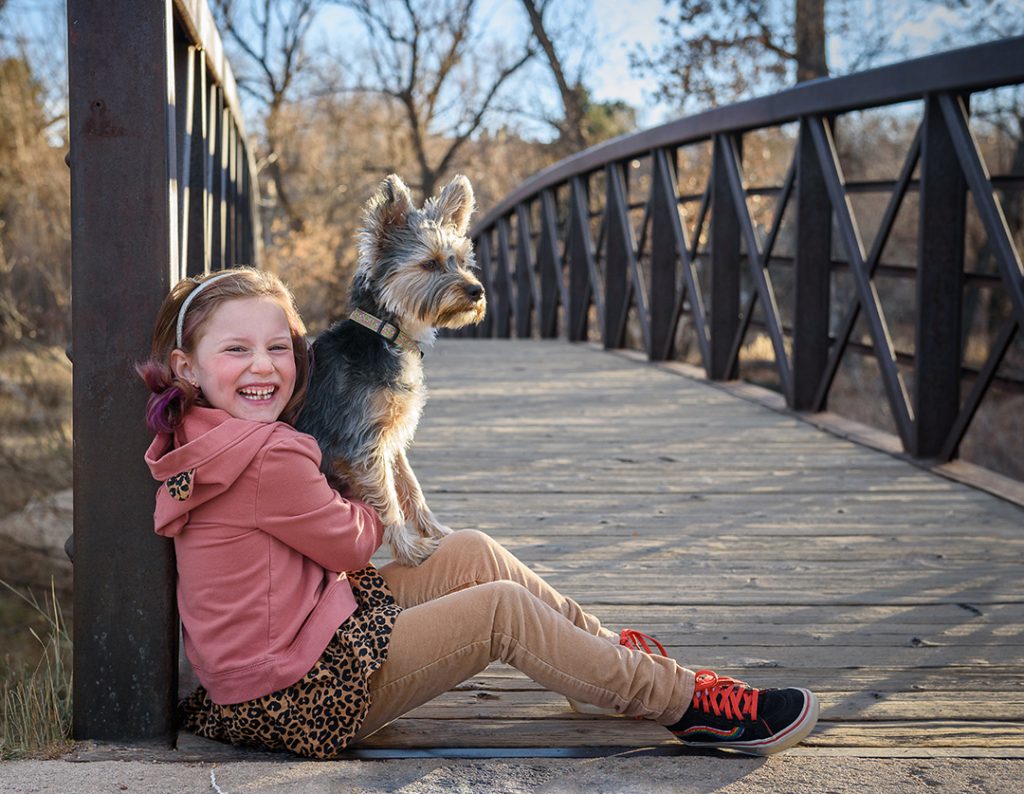 Picture of young child wearing a pink sweatshirt and her Terrier mix dog sitting on bridge