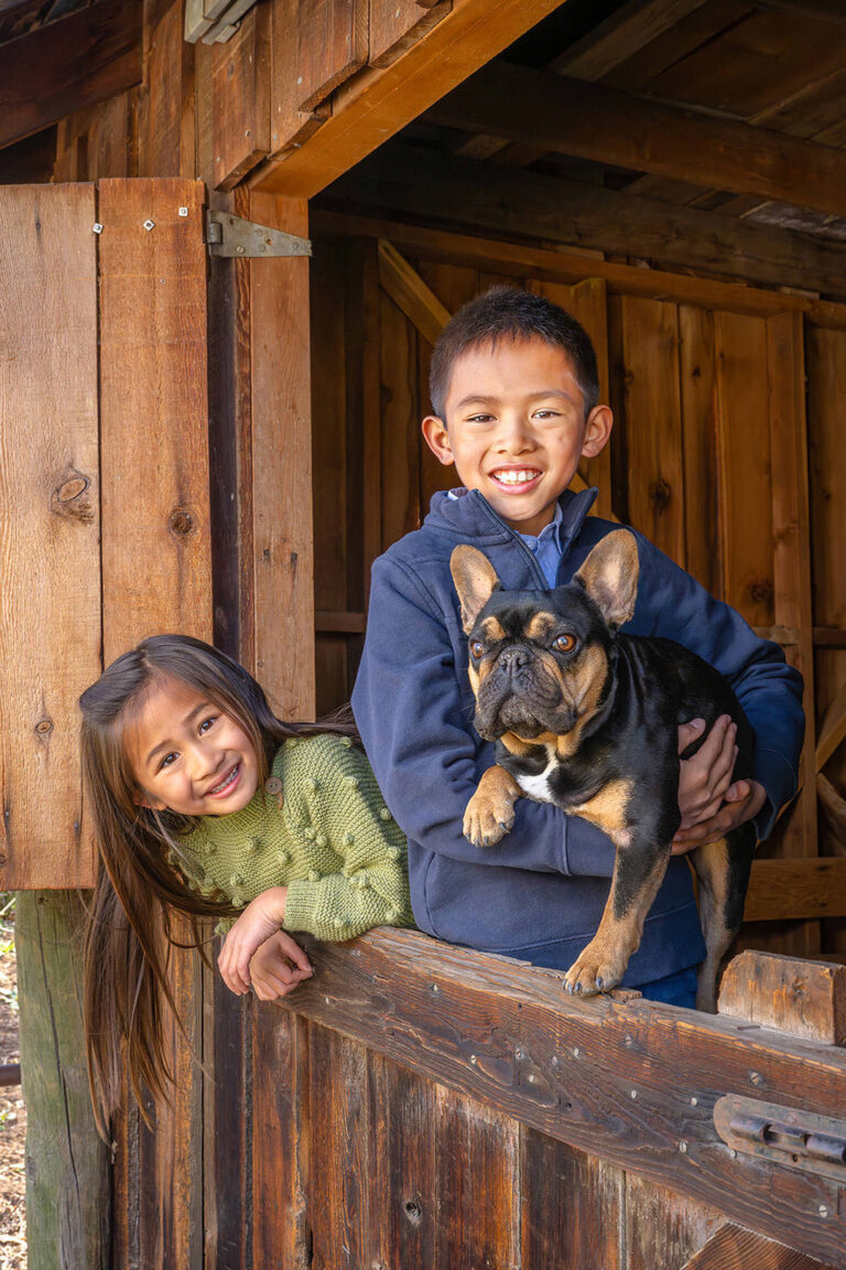 Two children posing in Dutch Door of barn and holding French Bulldog