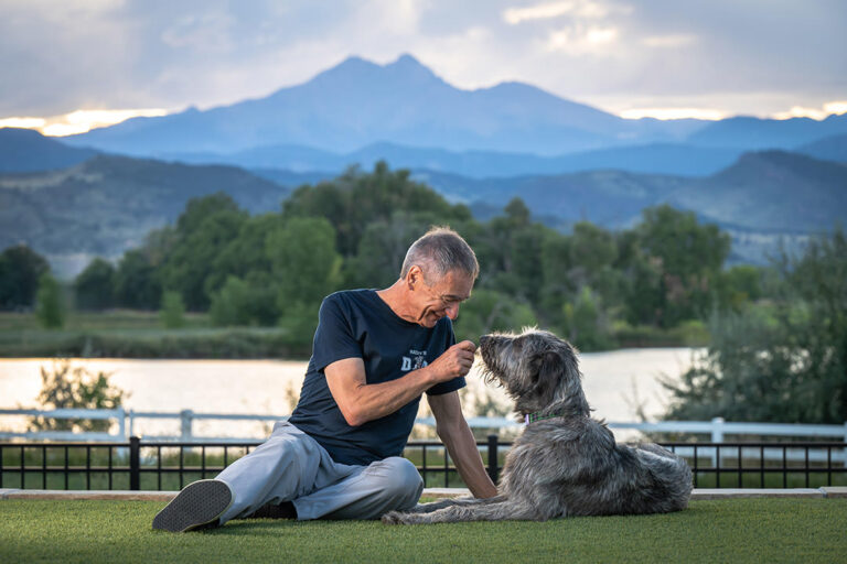Dog and Owner Photoshoot with mountains in background. Man and Irish Wolfhound sitting on grass with mountain and lake views in background