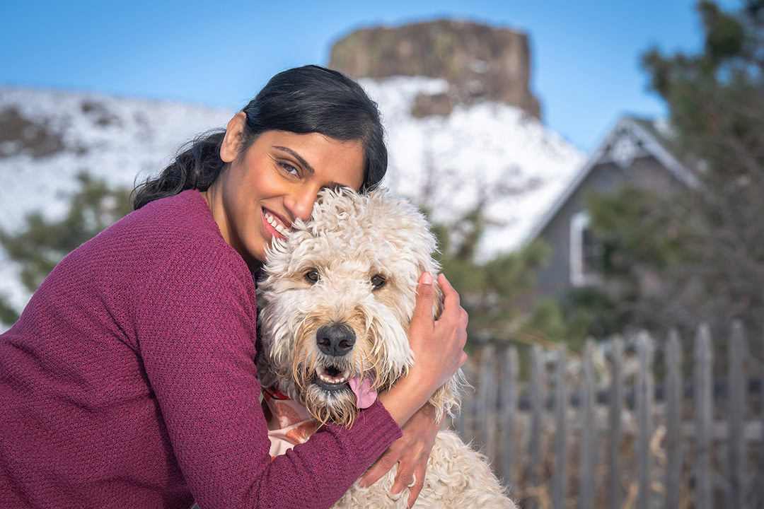 Woman in winter sweater hugging white goldendoodle dog with gray house and mountain in the background