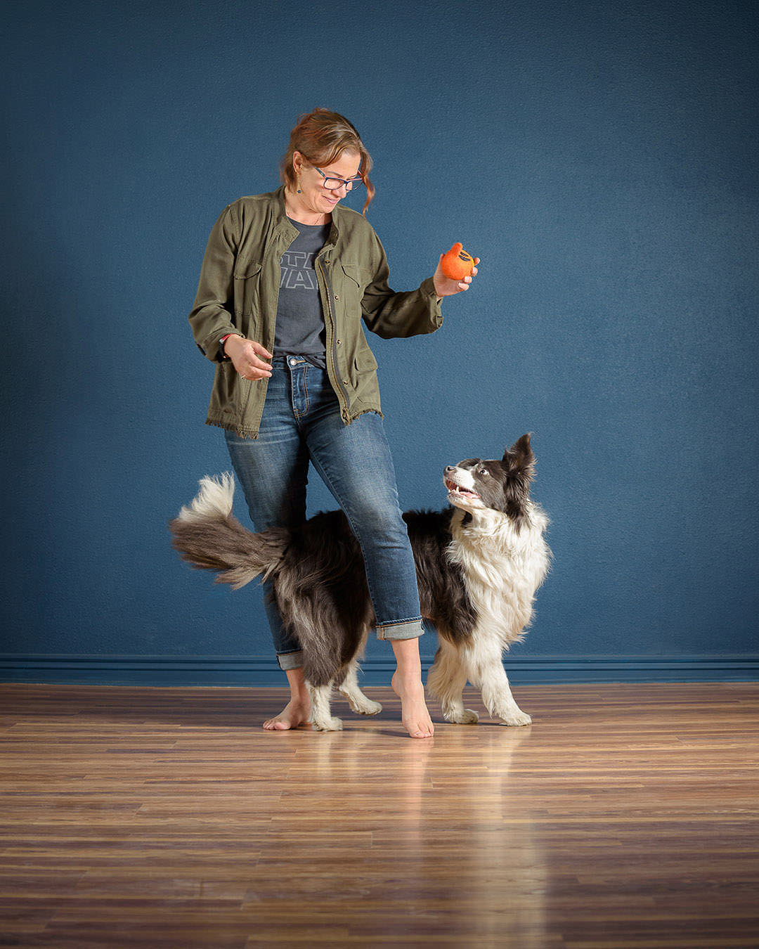 Border collie weaving in between woman's legs while looking at orange ball in her hand.