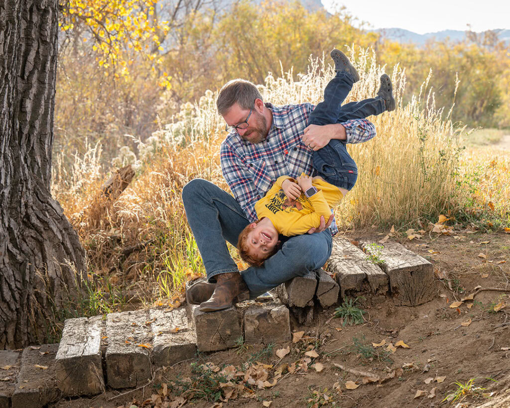 Dad and young son playing. Dad is seated and is holding son upside down. Son is laughing. Fall colors and mountain scenery behind them