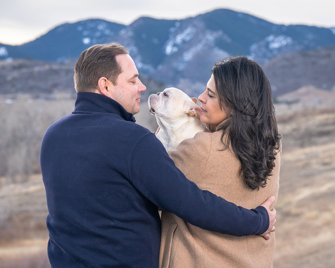 Couple photographed from behind with mountains in background. They are holding their while French bulldog who is looking lovingly at the man