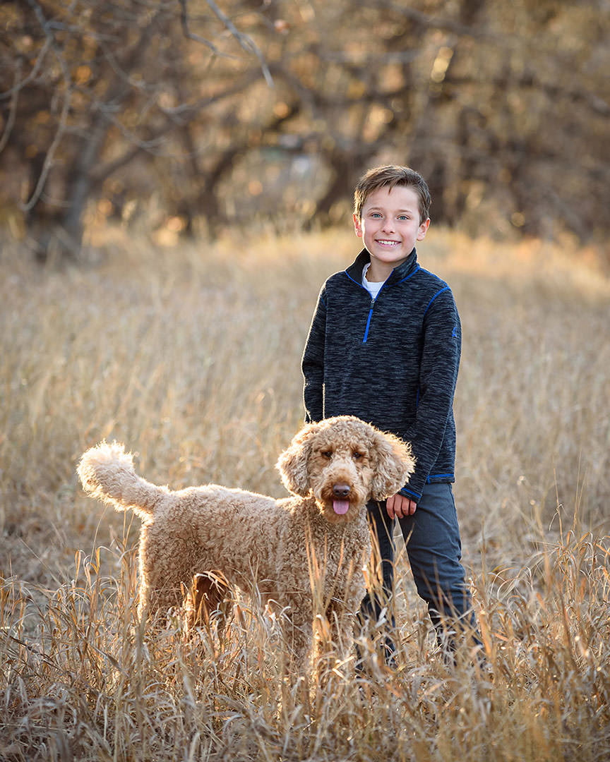 Young boy standing next to his goldendoodle dog in winter scenery with brown grasses and trees