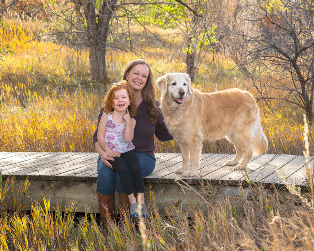 Mother and daughter sitting on boardwalk with their golden retriever on a fall day with yellow leaves on trees. Demonstrating mother and daughter photoshoot with dog