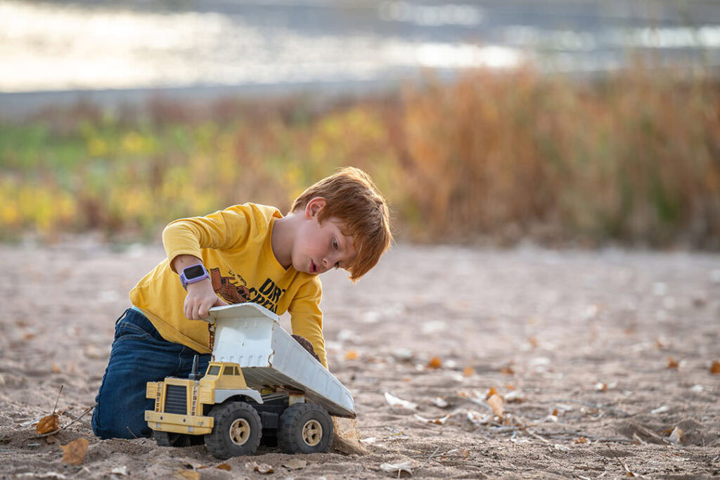 Young boy playing with a dump truck toy on beach with fall colors and lake behind him