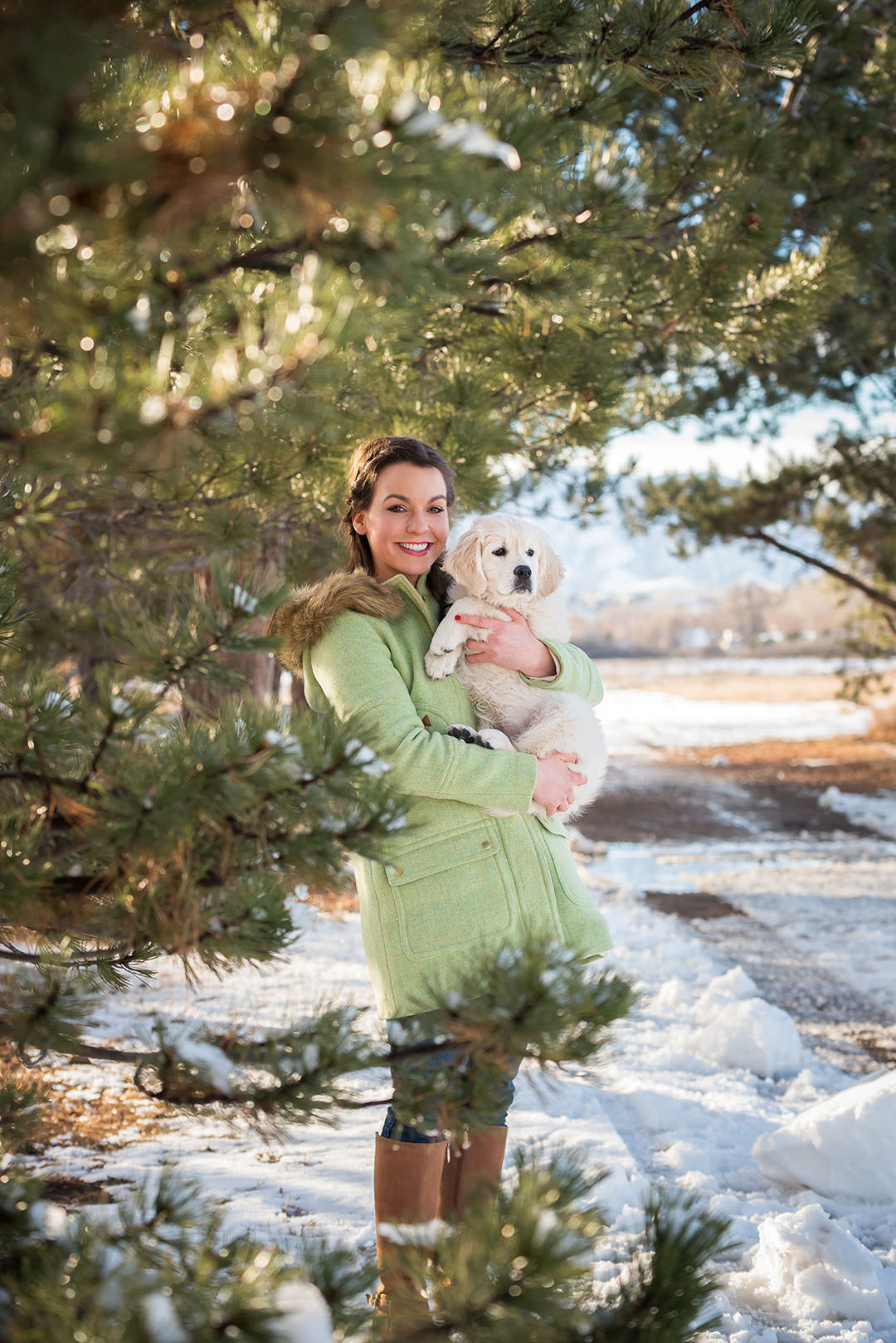 Woman holding golden retriever puppy. Framed by pine trees and snow is on ground.
